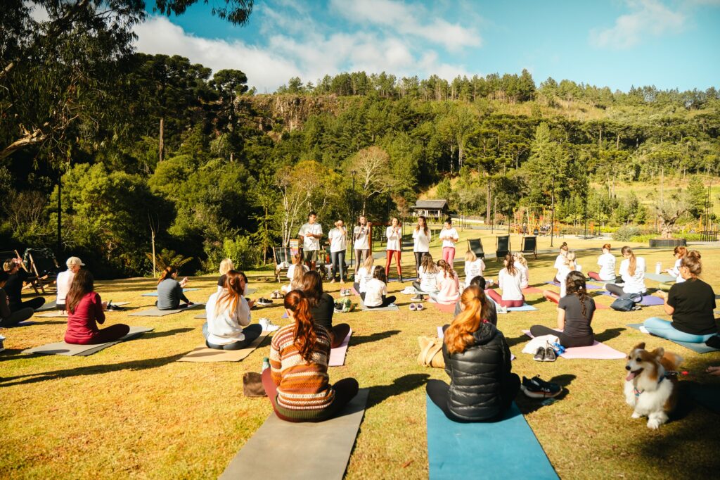 Foto horizontal mostra 26 pessoas sentadas sobre tapetes de EVA, dispostos em um grande gramado do Parque Paraíso das Araucárias. Elas estão de costas para foto. À frente delas estão seis pessoas - cinco mulheres e um homem, jovens. Todos usam camiseta branca. Uma das mulheres fala ao microfone. Ao fundo, aparecem muitas árvores, espreguiçadeiras e um playground. No canto direito inferior da foto, aparece um cachorro com pelos brancos e beges com a língua para fora.