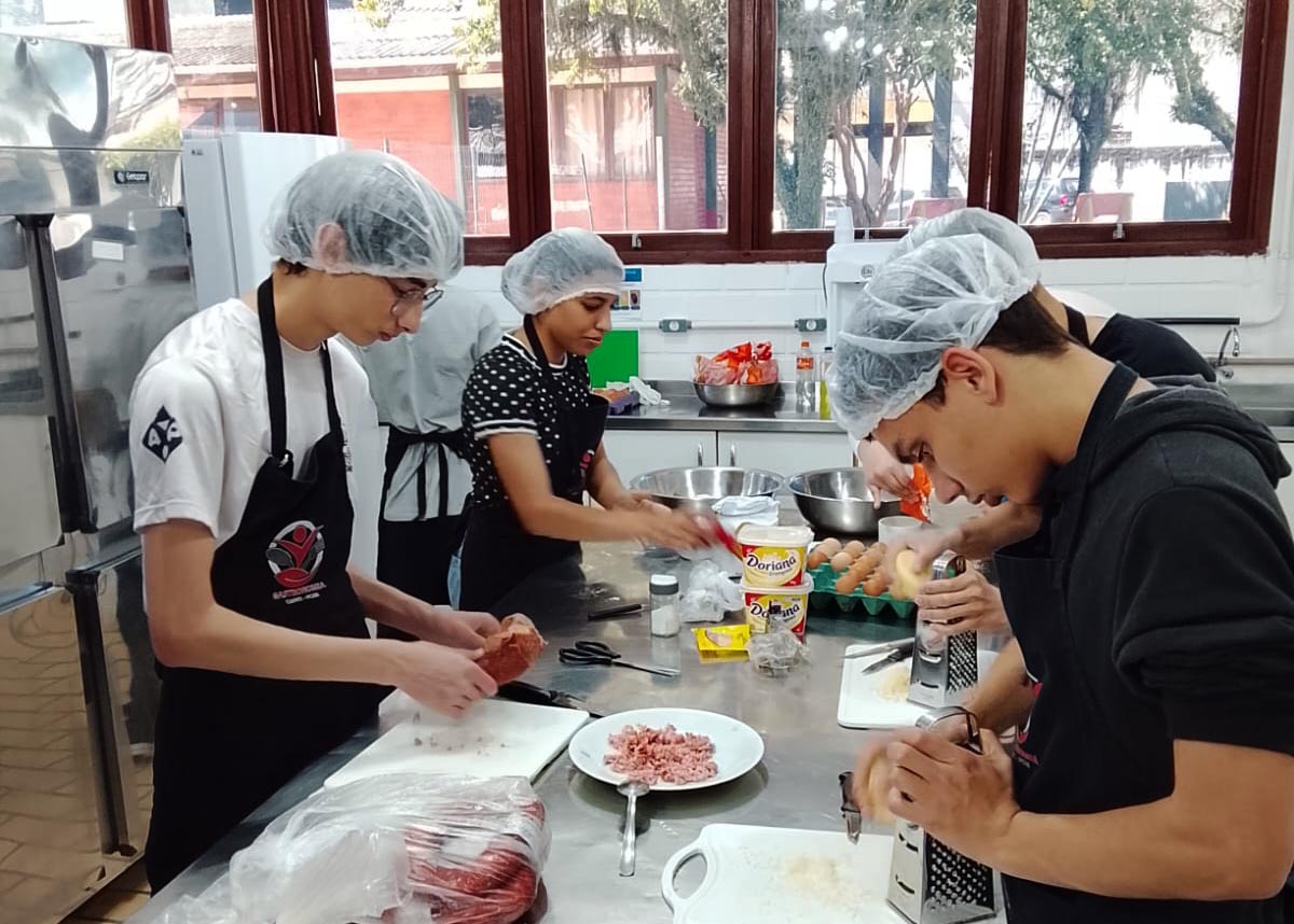Grupo de jovens em cozinha industrial, manipulando alimentos sobre mesa de inox com trajes de cozinha.