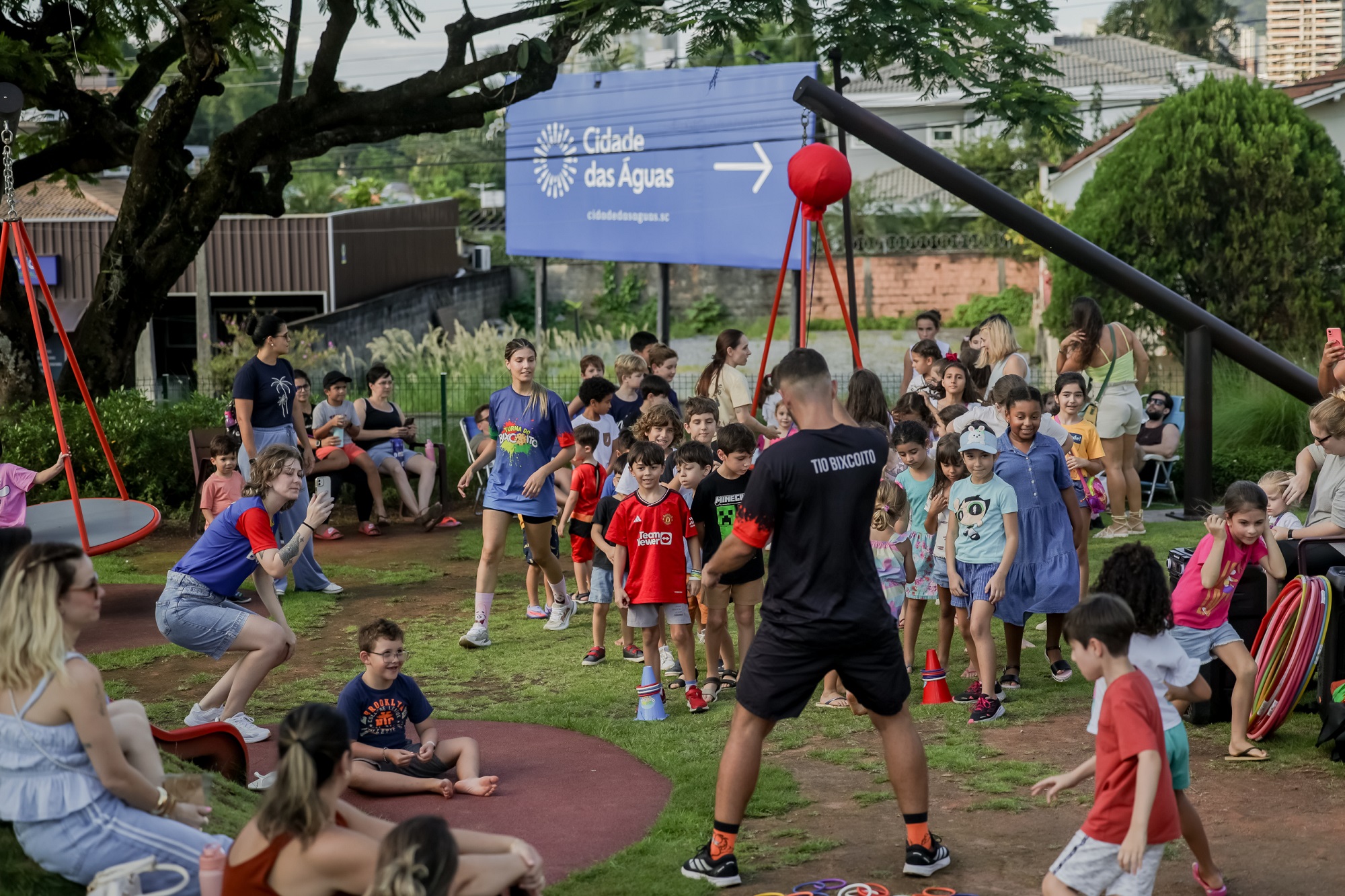Foto horizontal mostra dezenas de crianças de diferentes idades e características físicas e alguns adultos na Praça Cidade das Águas. Elas estão sobre uma área gramada. Parte delas está em pé, outras correm pelo espaço e algumas estão sentadas. Há adultos com camisetas de recreadores que interagem com elas. No entorno aparecem algumas árvores e uma grande placa azul com a logomarca da Cidade das Águas.